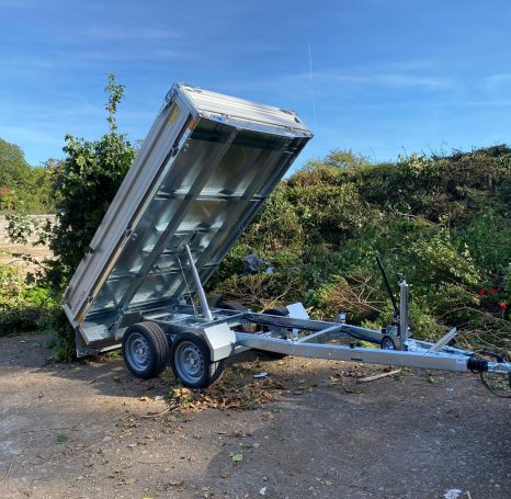 Ein kippbarer Anhänger steht auf unbefestigtem Boden, umgeben von Gestrüpp.
