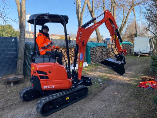 Frau in orangefarbener Arbeitsjacke sitzt auf einem kleinen, orangenen Bagger.